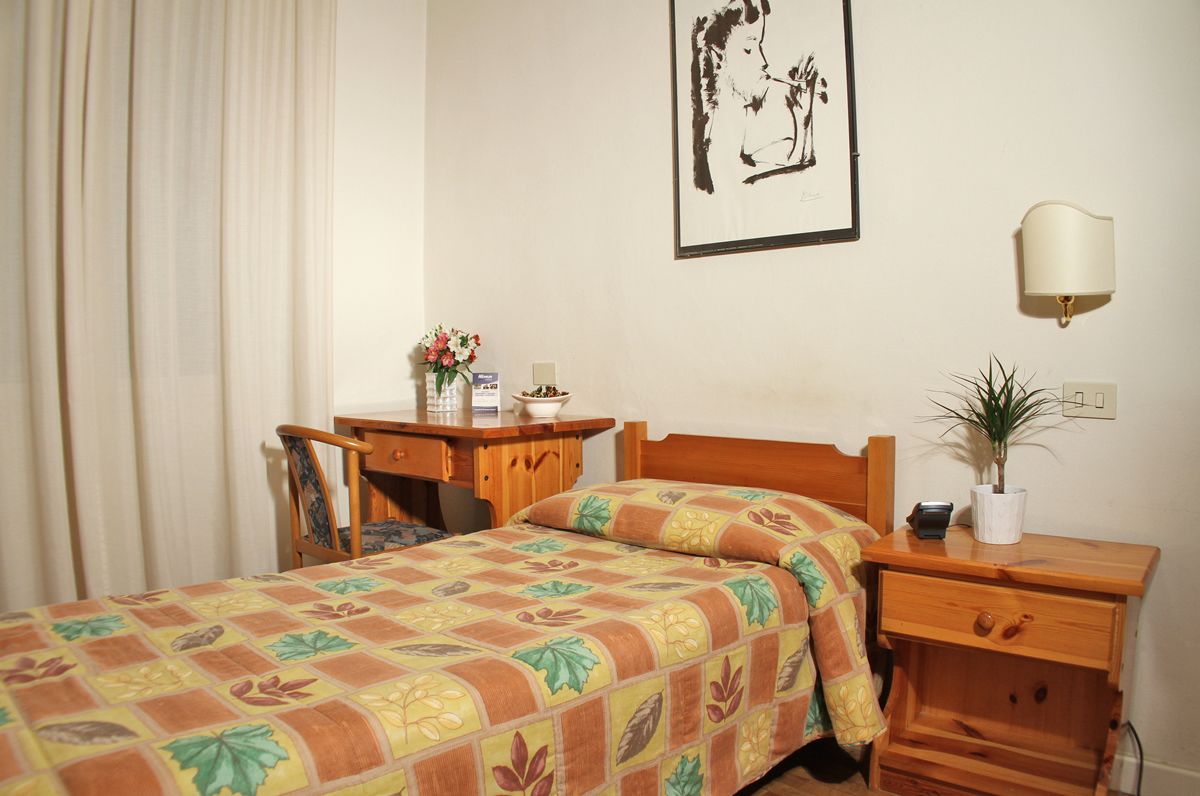 A bedroom with a bed, a desk, a chair, a bedside table with a lamp, and a framed picture on the wall.