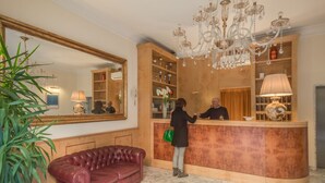 A reception area with a wooden reception desk, a large mirror, a chandelier, and a red leather couch. - Hotel La Pace (Pontedera)