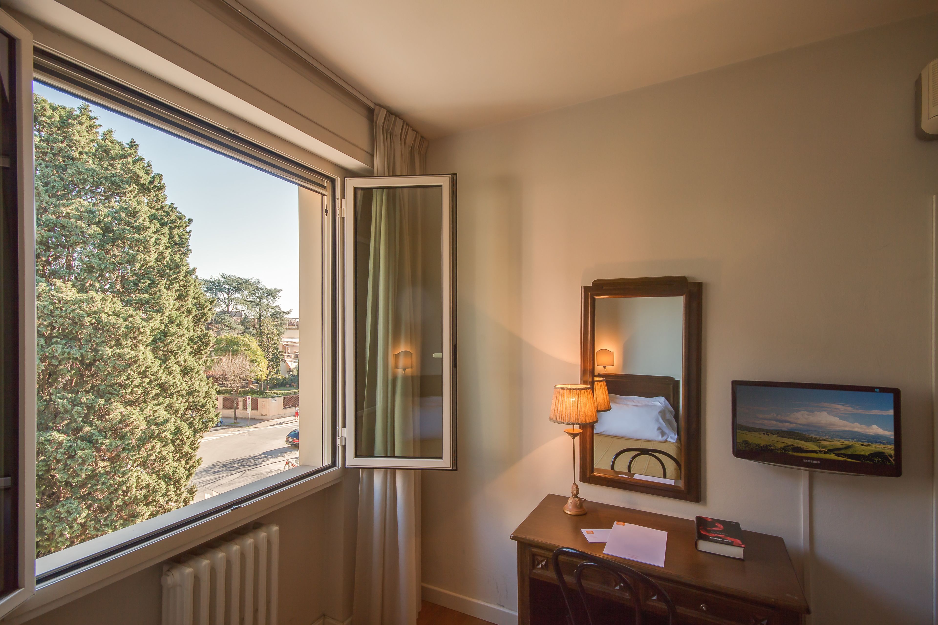 Ein Hotelzimmer mit großem Fenster, Schreibtisch mit Lampe, Spiegel und Flachbildfernseher.