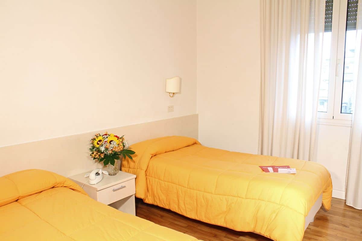 Une chambre d’hôtel avec deux lits, une table de chevet ornée d’un vase de fleurs, une lampe et une fenêtre avec des rideaux.