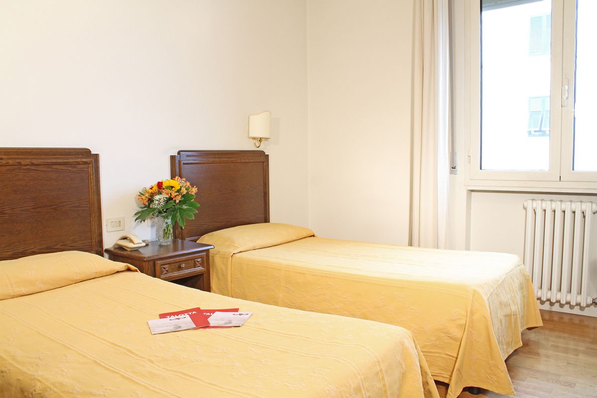 A hotel room with two beds, a nightstand with a lamp, a vase with flowers, and a window with a view of buildings.