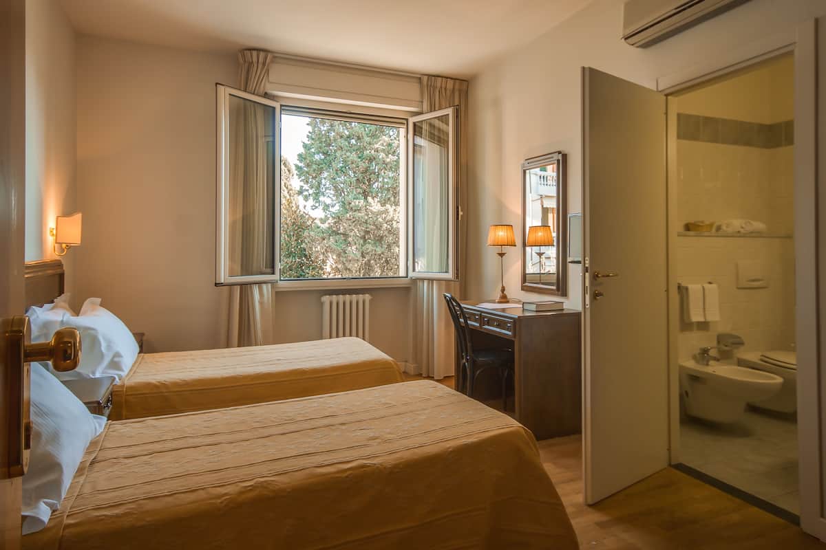 Une chambre d’hôtel avec deux lits, un bureau avec une chaise, une fenêtre avec des rideaux, une salle de bain avec une douche et un miroir.