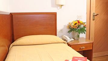 Una camera d'albergo con una testiera in legno, un letto con copripiumino giallo, un comodino con una lampada, un vaso con fiori e un libretto rosso etichettato 'TALOTTA'.