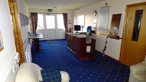 A hotel room with a blue carpet, a white armchair, a desk with a computer, a wooden desk, a clock, and a door with a 'No Smoking' sign. - Orkneylodge (Orphir)