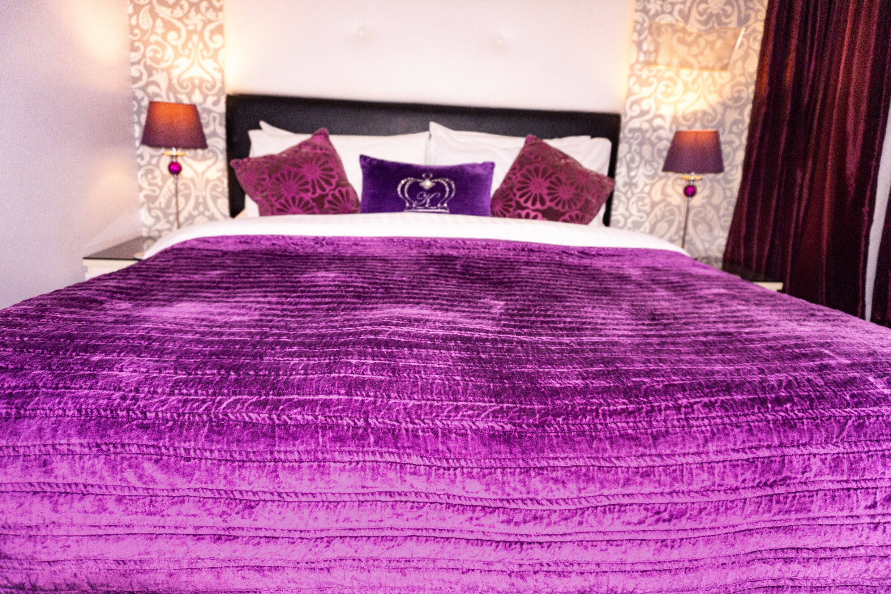 A bed with a purple bedspread and decorative pillows, two bedside lamps, and patterned wallpaper.