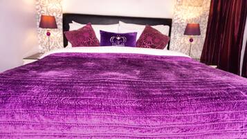 A bed with a purple bedspread and decorative pillows, two bedside lamps, and patterned wallpaper.