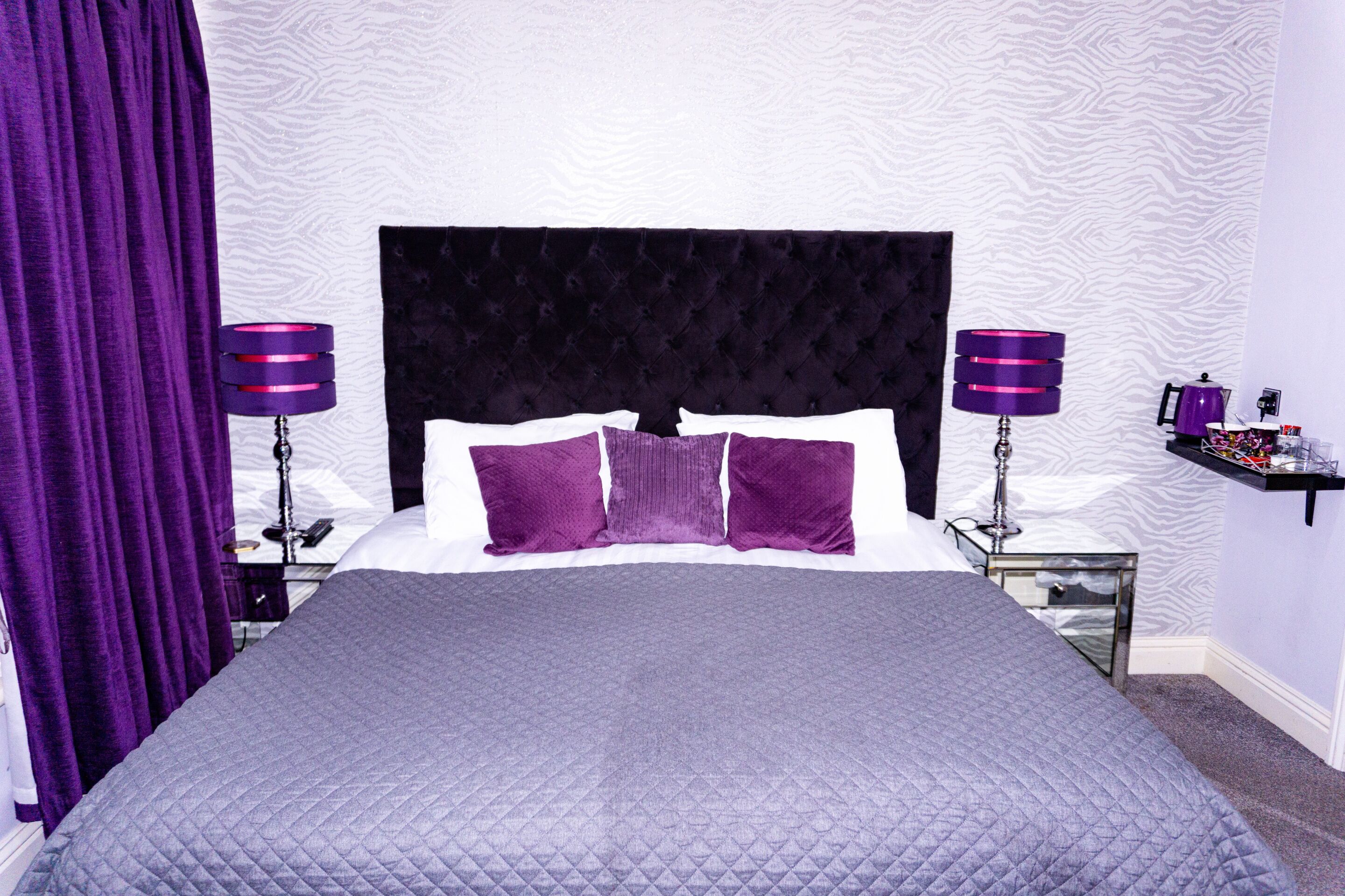 A neatly made bed with a dark headboard, two purple lamps on either side, and a small shelf with items on the nightstand.