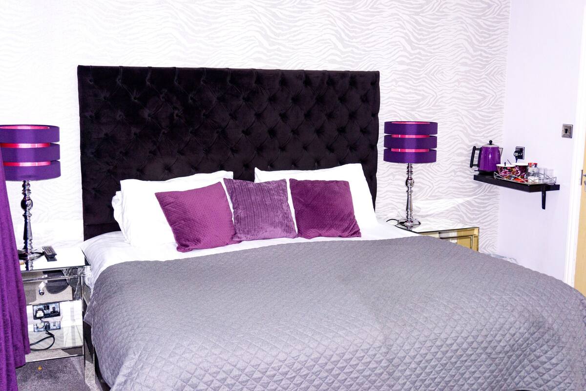 A large bed with a tufted headboard, two bedside tables with lamps, and a small shelf with toiletries.