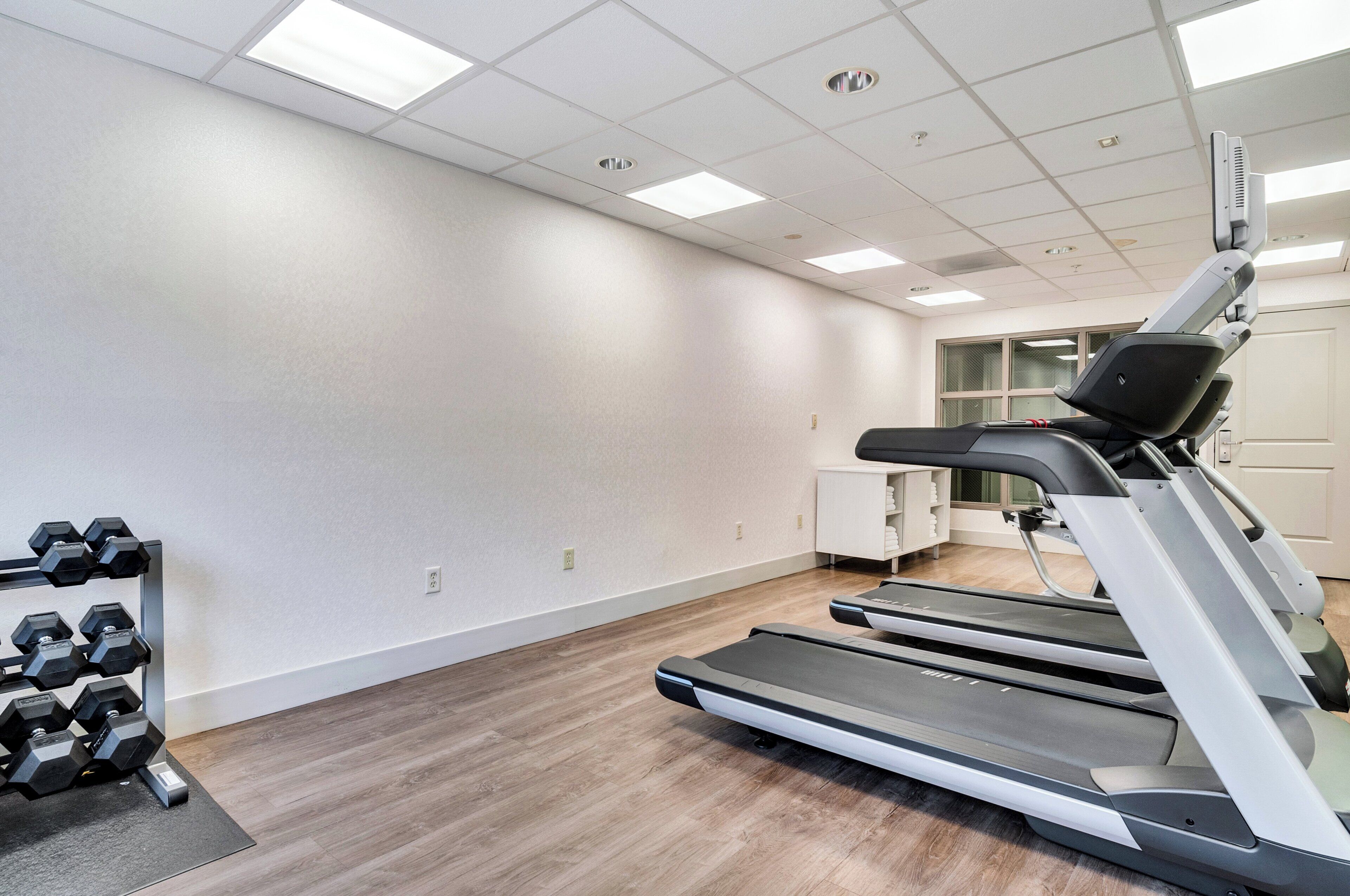 A gym with treadmills, a dumbbell rack, and a window with blinds.