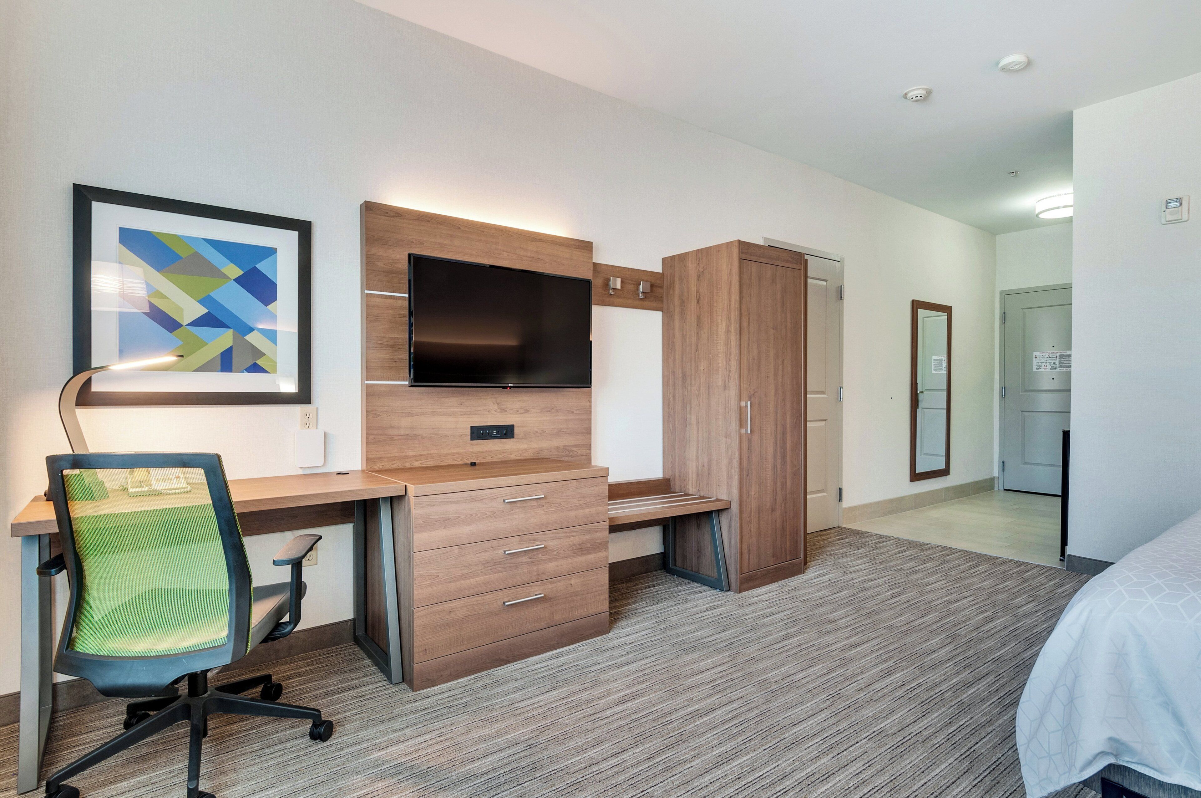 A modern hotel room with a wooden desk, a chair, a flat-screen TV, a wardrobe, and a bed.