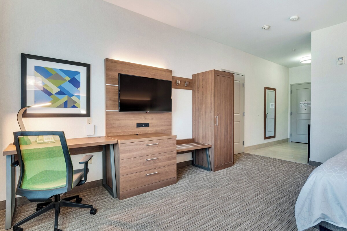 A modern hotel room with a wooden desk, a chair, a flat-screen TV, a wardrobe, and a bed.