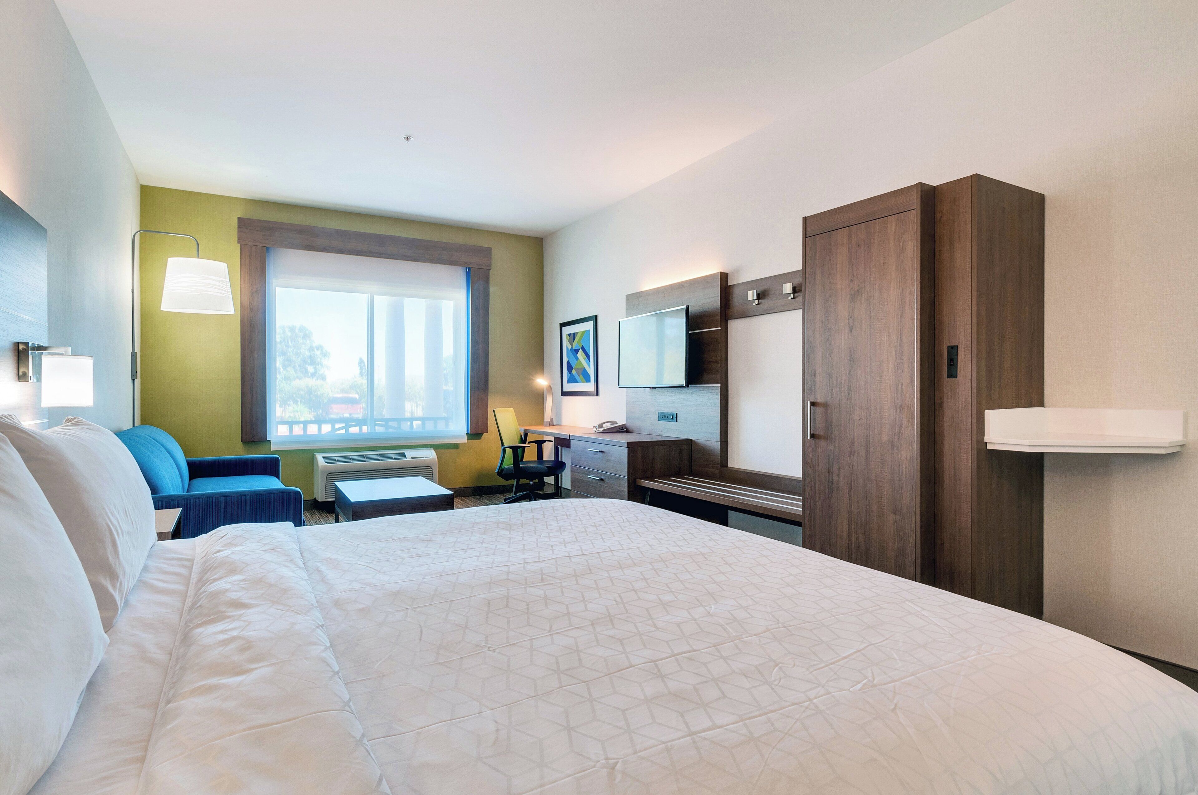 Une chambre d’hôtel avec un grand lit, un bureau avec une chaise, une petite table, une lampe, une télévision et une armoire.