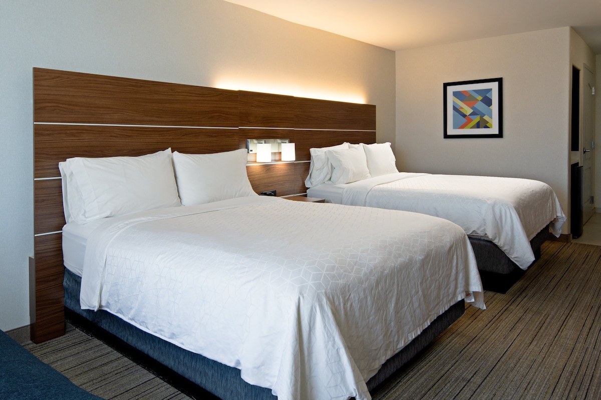 A hotel room with two beds, a wooden headboard, a nightstand with a lamp, and a framed abstract artwork on the wall.