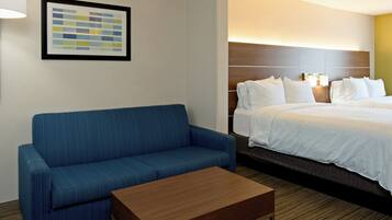 A hotel room with a blue sofa, a wooden coffee table, a bed with white bedding, and a framed picture on the wall.