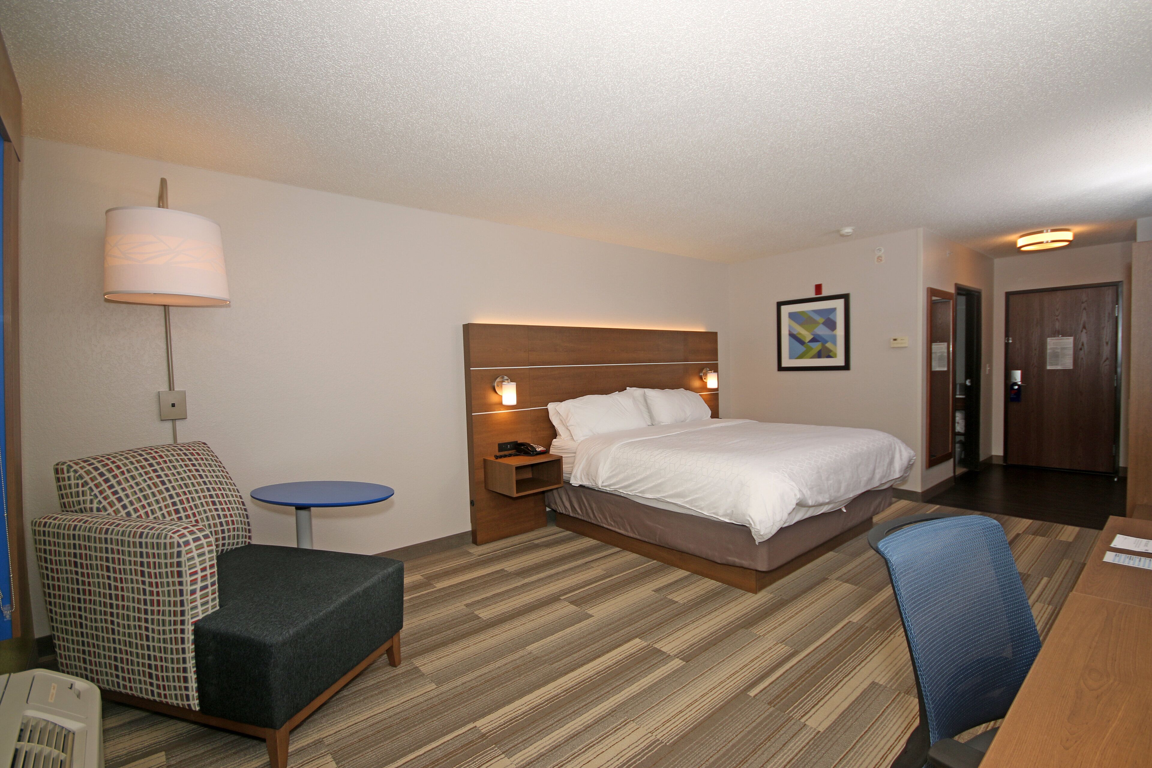 A hotel room with a bed, a chair, a small table, and a lamp.