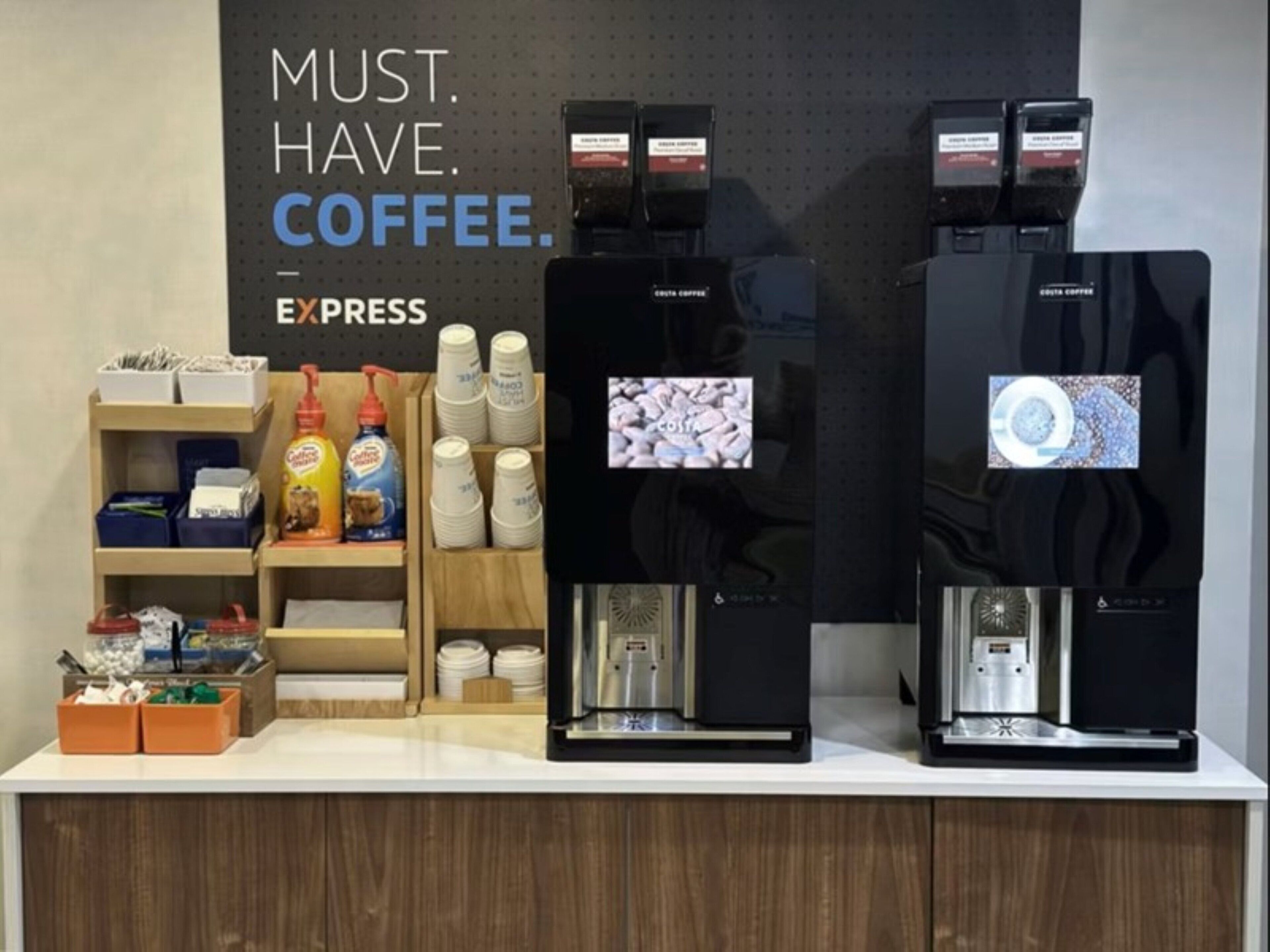A coffee station with two espresso machines, a sign reading "MUST. HAVE. COFFEE.", and various coffee pods and cups.