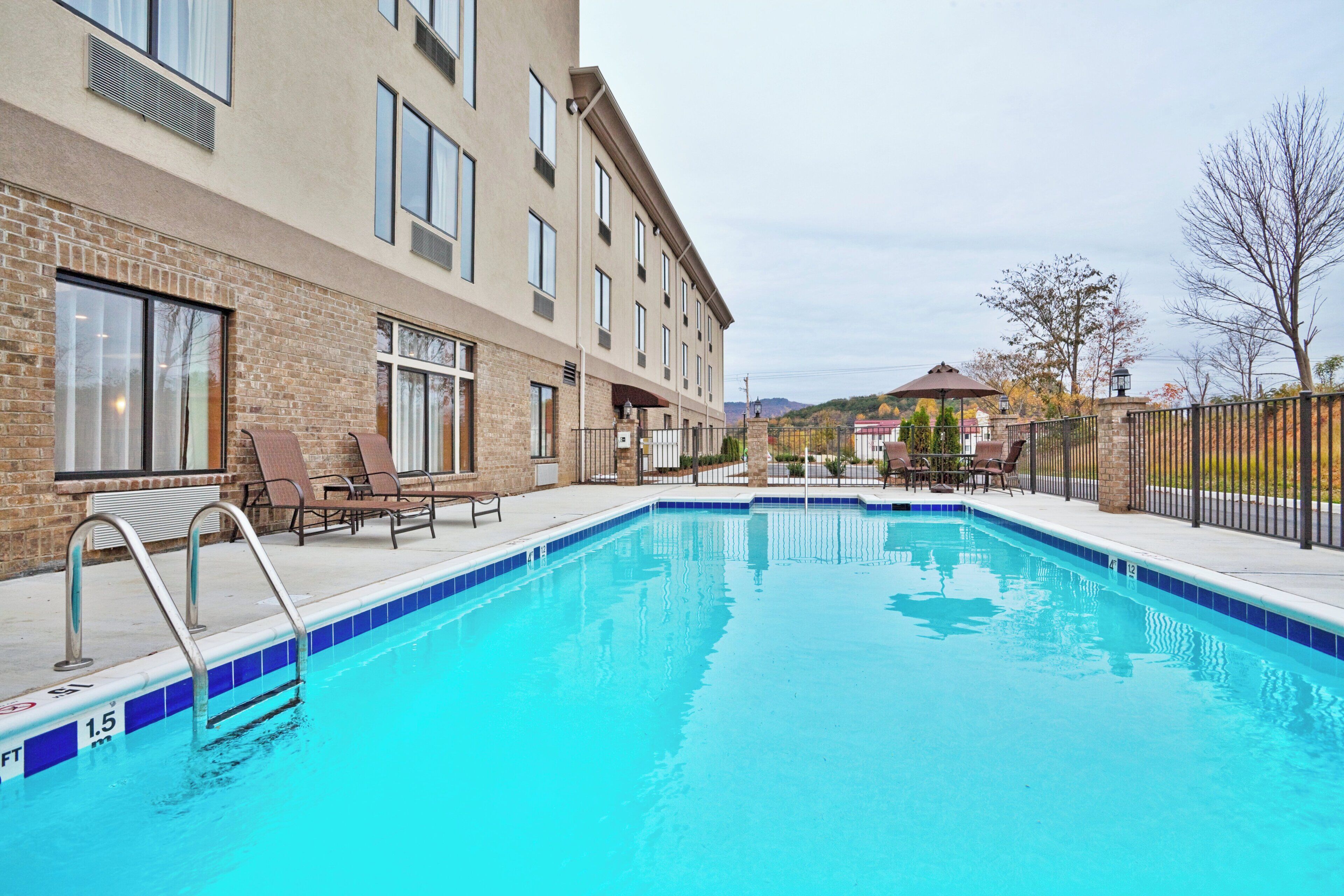 A swimming pool with a clear blue water edge, surrounded by a brick building with multiple windows and a covered seating area with chairs.