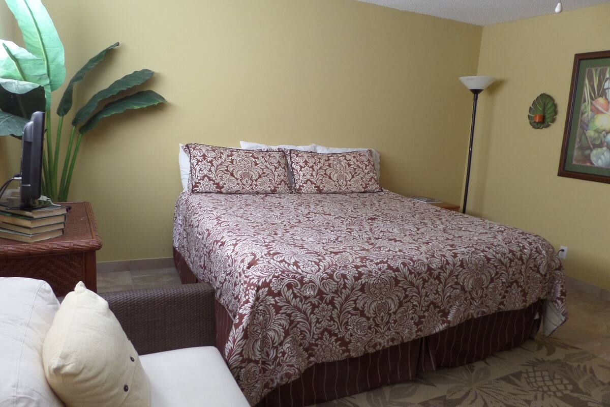 A bedroom with a bed, a nightstand, a lamp, and a large plant.