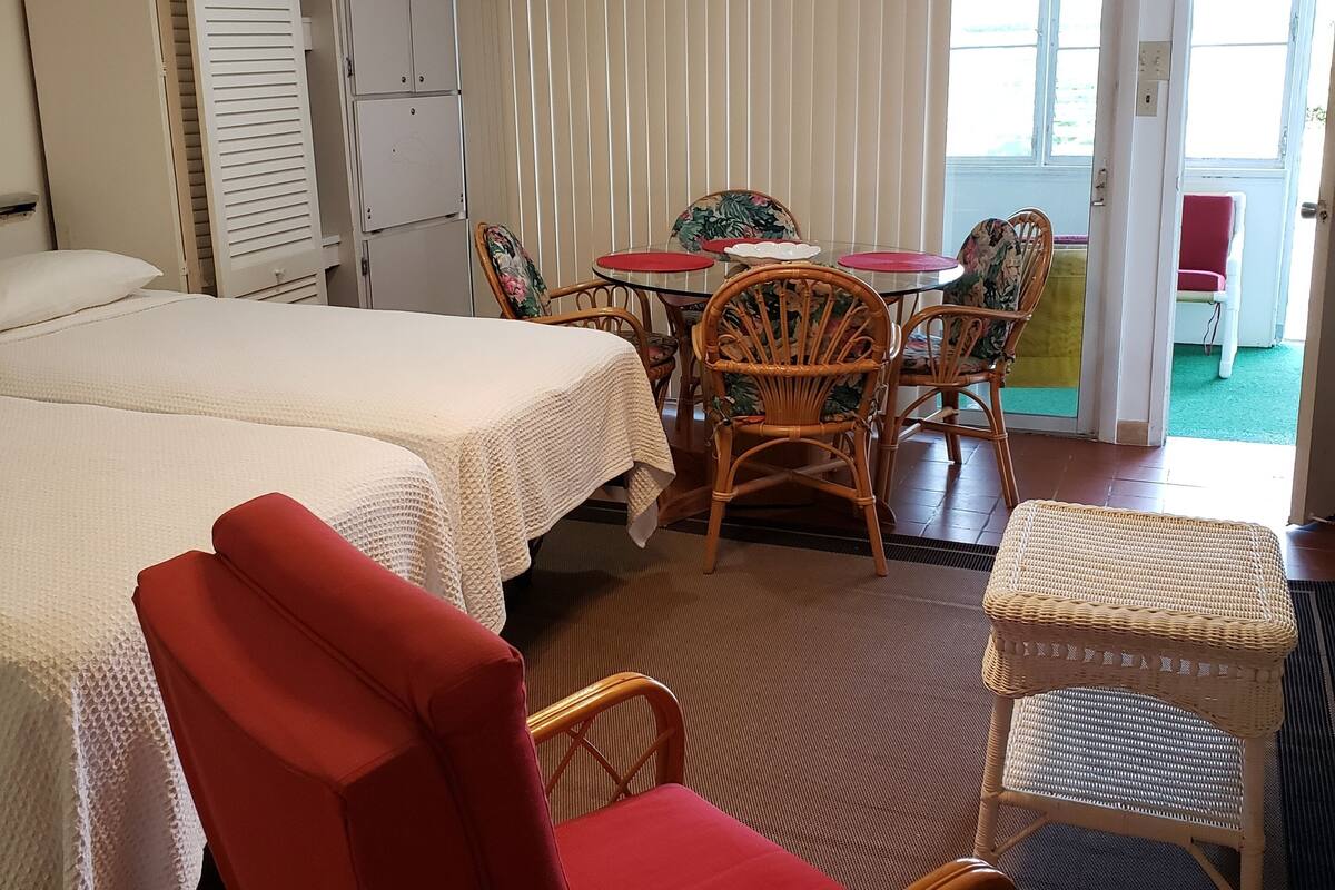 A hotel room with two beds, a dining table with chairs, a ceiling fan, and a view of the outside.