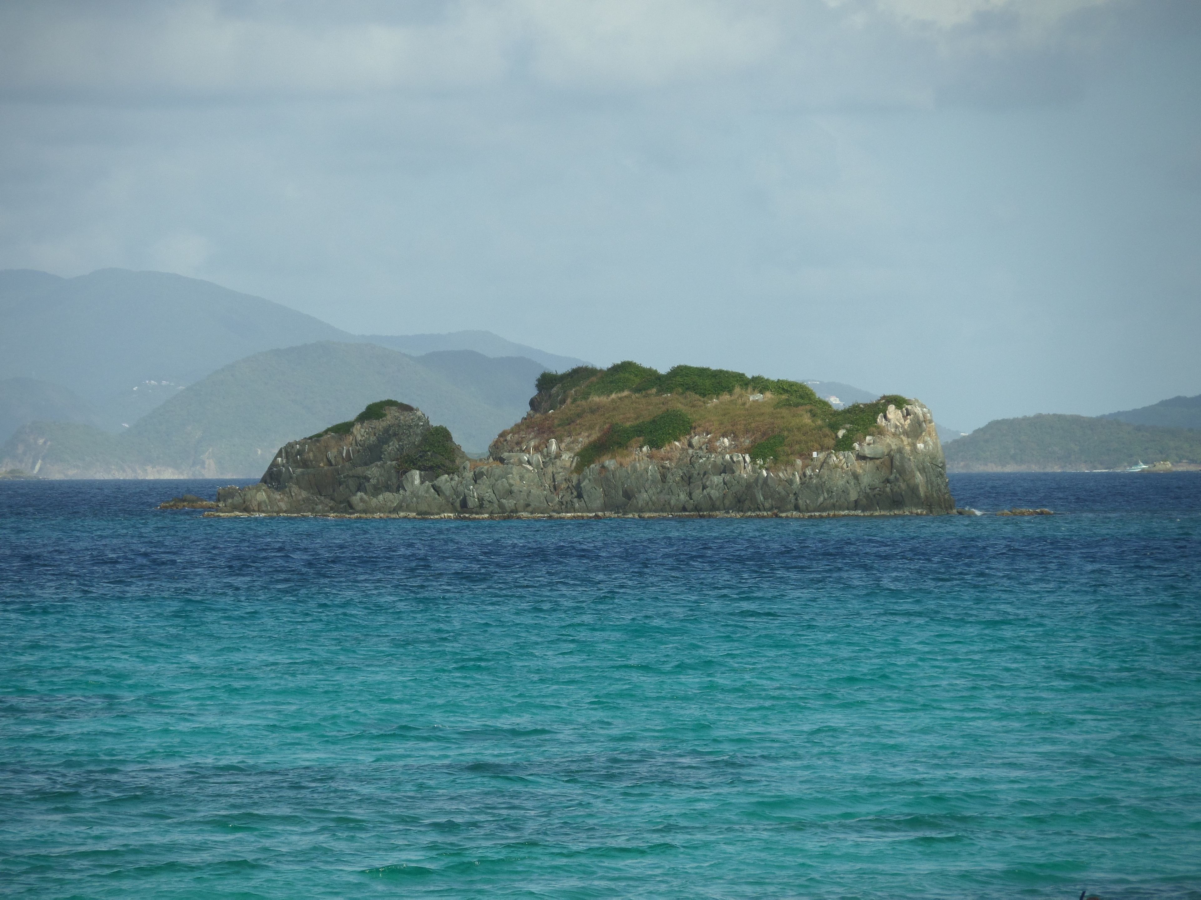 A rocky island with greenery in the middle of a calm sea, surrounded by mountains.