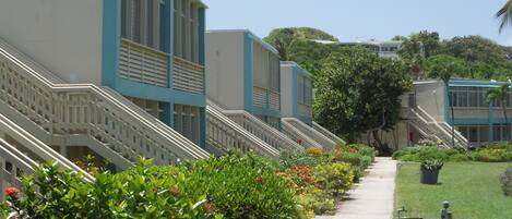 A row of hotel buildings with multiple floors and balconies, a pathway lined with colorful flowers, and a grassy area with a trash bin.