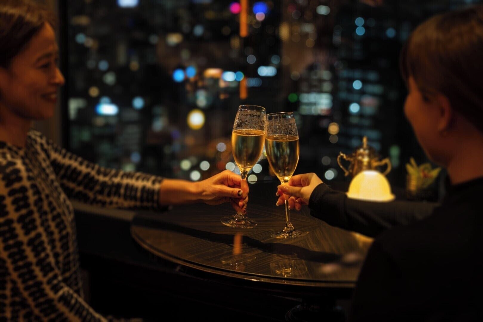 Two people toasting with champagne glasses at a table with a city view in the background.