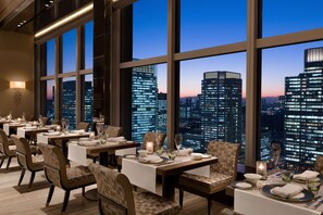 Un restaurante con mesas dispuestas para comer, grandes ventanales que ofrecen vista al horizonte de la ciudad y un diseño interior moderno.