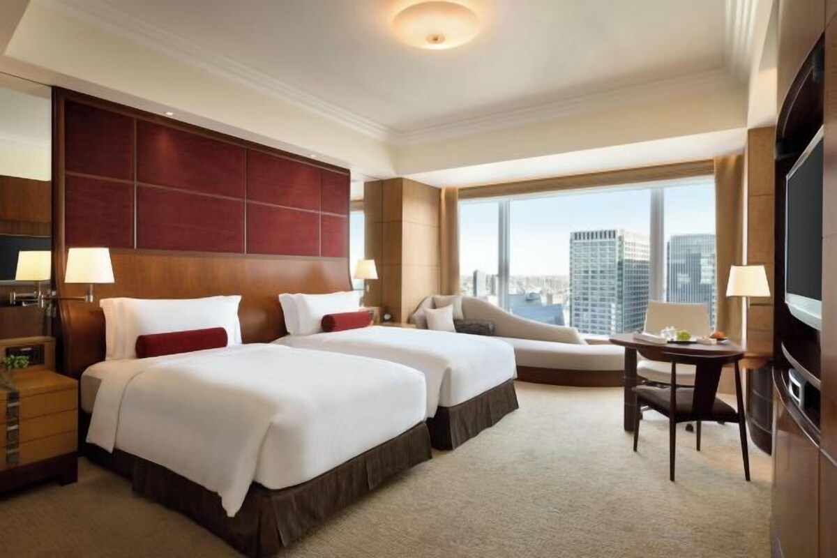 A hotel room with two beds, a sofa, a TV, and a large window with a city view.