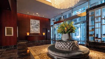 A lobby with a marble floor, a large chandelier, a reception desk, a wall-mounted artwork, and a flower arrangement on a cushioned stool.