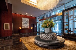 A lobby with a marble floor, a large chandelier, a reception desk, a wall-mounted artwork, and a flower arrangement on a cushioned stool. - Shangri-La Tokyo (Tokyo)