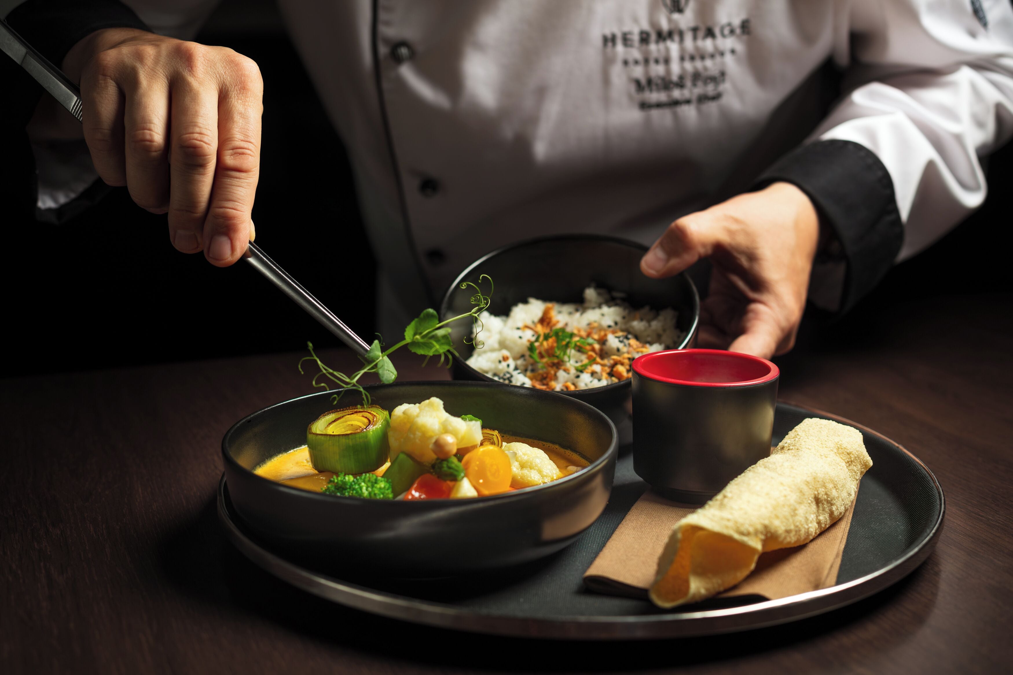 Un chef présente un plat composé de riz, de légumes et d’une préparation roulée, le tout servi sur un plateau.