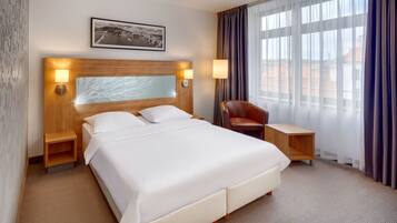 A hotel room with a large bed, two bedside tables with lamps, a wooden headboard, a framed picture on the wall, a window with curtains, and a leather chair.