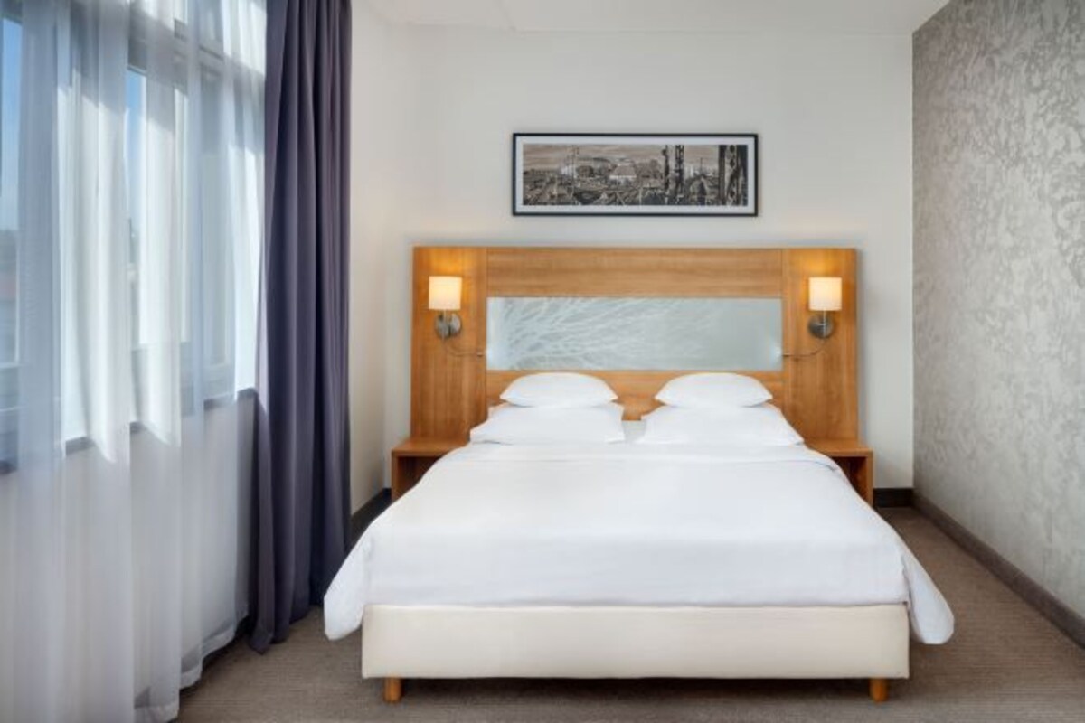 Habitación de hotel con cama, cabecero con lámparas de pared y un cuadro enmarcado, además de cortinas translúcidas.