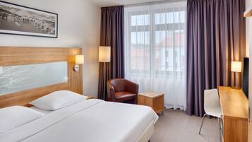 Ein Hotelzimmer mit einem großen Bett, einem Schreibtisch, einem Stuhl und einem fensterbelaubten Fenster.