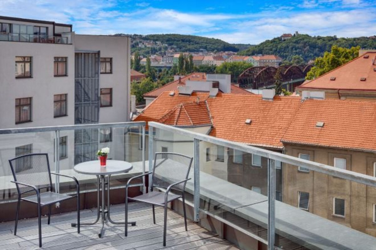 Un balcón con una mesa y sillas, con vistas a un paisaje urbano con edificios de tejados rojos y una colina al fondo.