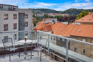 Varanda com mesa e cadeiras, vista para um horizonte urbano com edifícios de telhado vermelho e uma colina ao fundo.