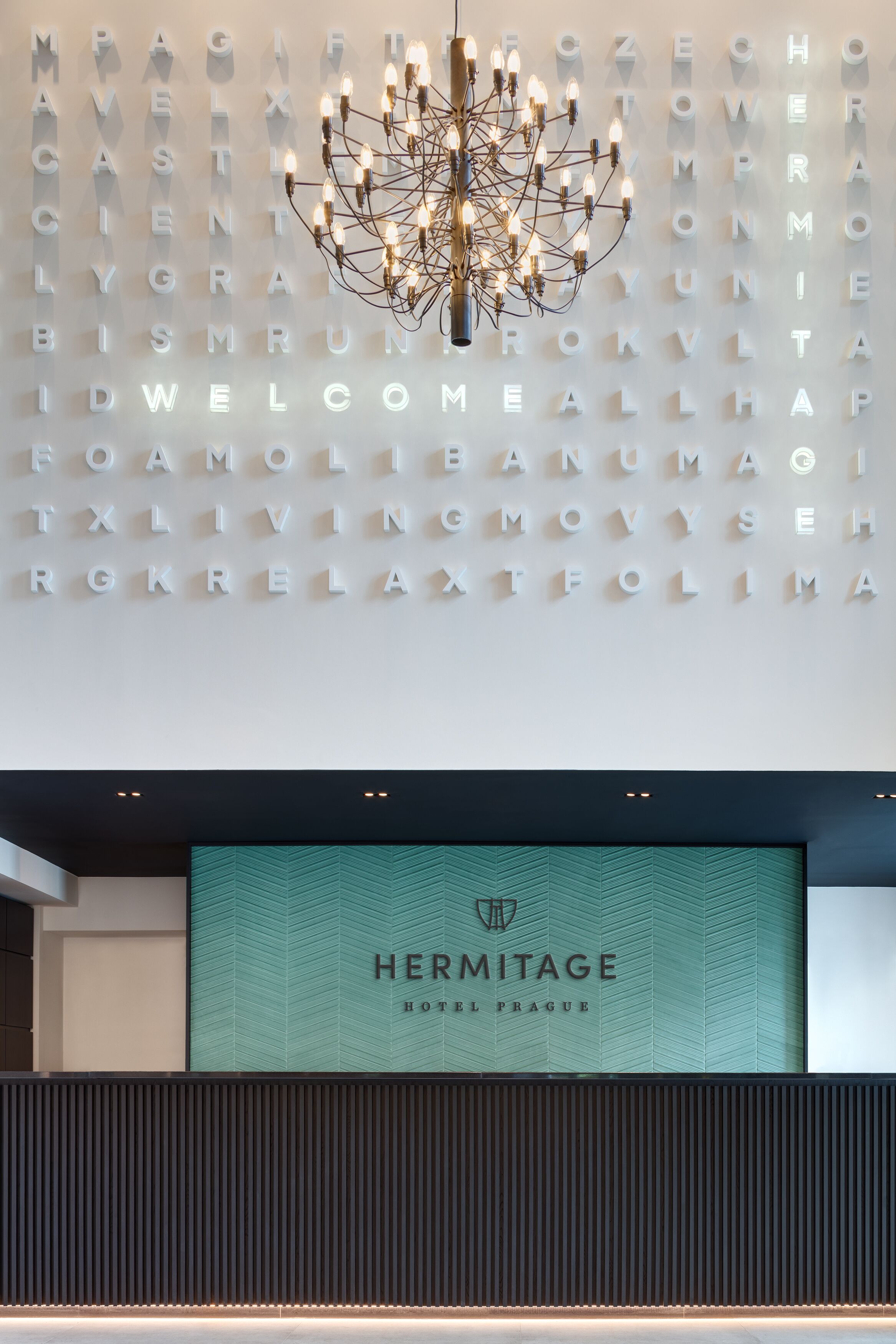 Recepção moderna de hotel com um candelabro, uma parede de letras iluminadas e uma tela grande exibindo o nome do hotel.