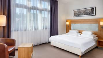 Habitación de hotel con cama, mesita de noche de madera, una pequeña mesa, una silla y un ventanal con cortinas translúcidas.