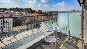 A balcony with a glass railing, a table with a chair, and a view of a cityscape. - Hermitage Hotel Prague (Prague)