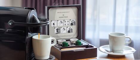 A coffee maker with a box of coffee pods, two white cups, and a plate with a spoon on a wooden table.