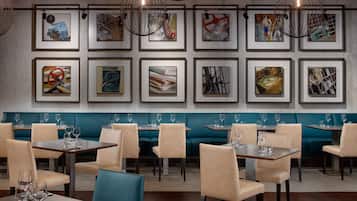 Un restaurante con asientos de tapicería azul verdoso, sillas color beige y cuadros en la pared.