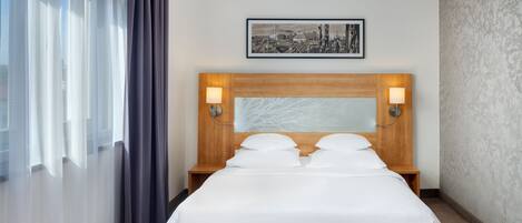 Una cama bien tendida con sábanas blancas, un cabecero de madera y dos lámparas de lectura fijadas a la pared.