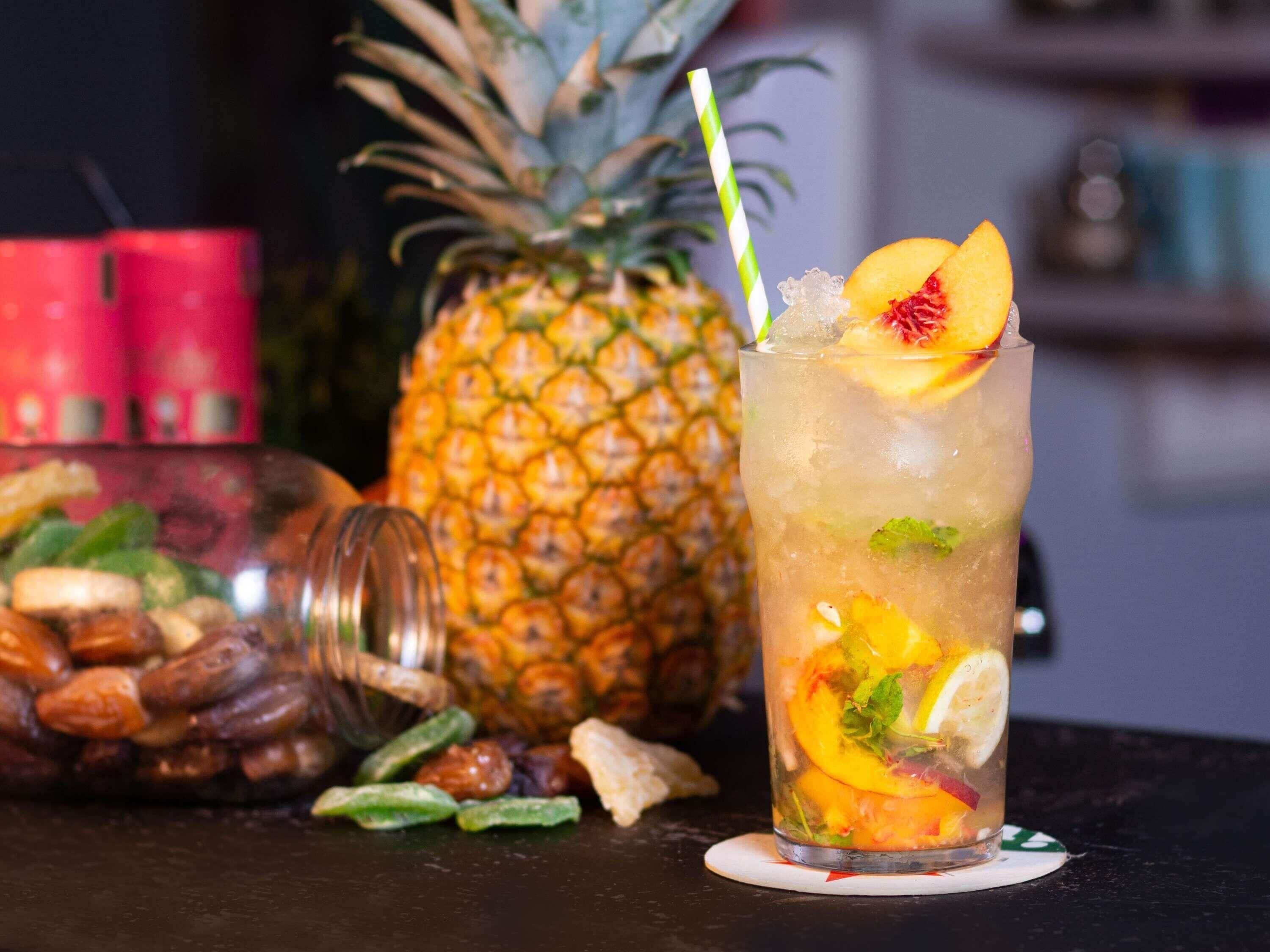 Un vaso de bebida con frutas y menta, una piña y un tarro de nueces sobre una mesa.