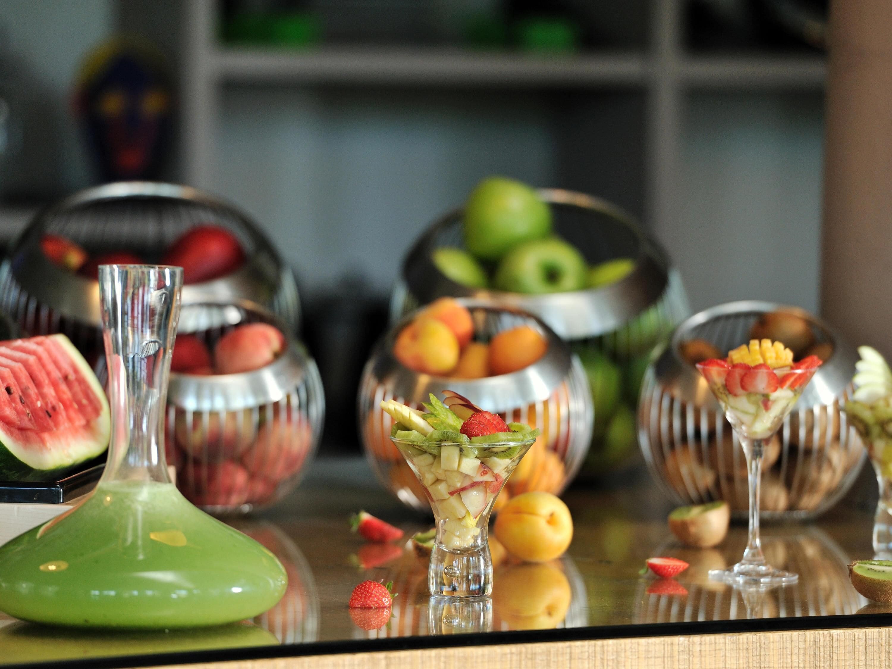 A table with fruit bowls, a green glass container, and a clear glass with a fruit garnish.