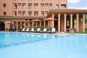 A swimming pool with lounge chairs and umbrellas, surrounded by a building with multiple balconies and a green roof.
