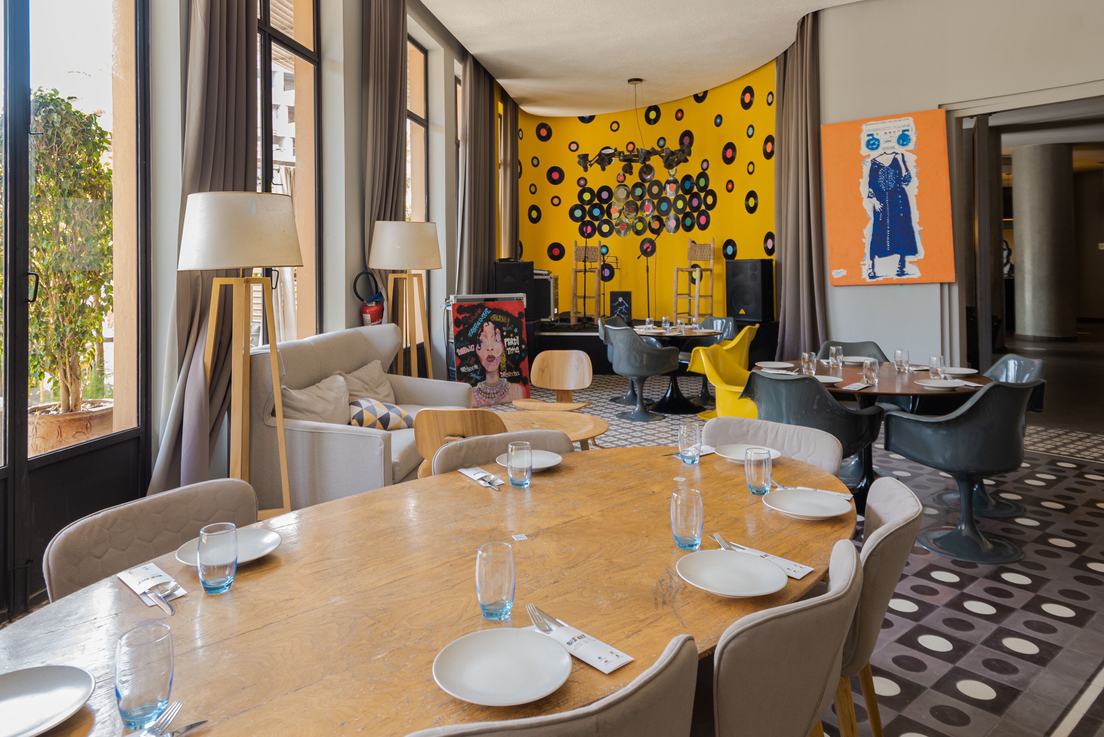 Un comedor con una mesa de madera dispuesta para cuatro personas, rodeada de sillas. Hay dos lámparas, un sofá y una mesa de centro. La habitación tiene una pared amarilla con diseños circulares y un cuadro de una mujer.