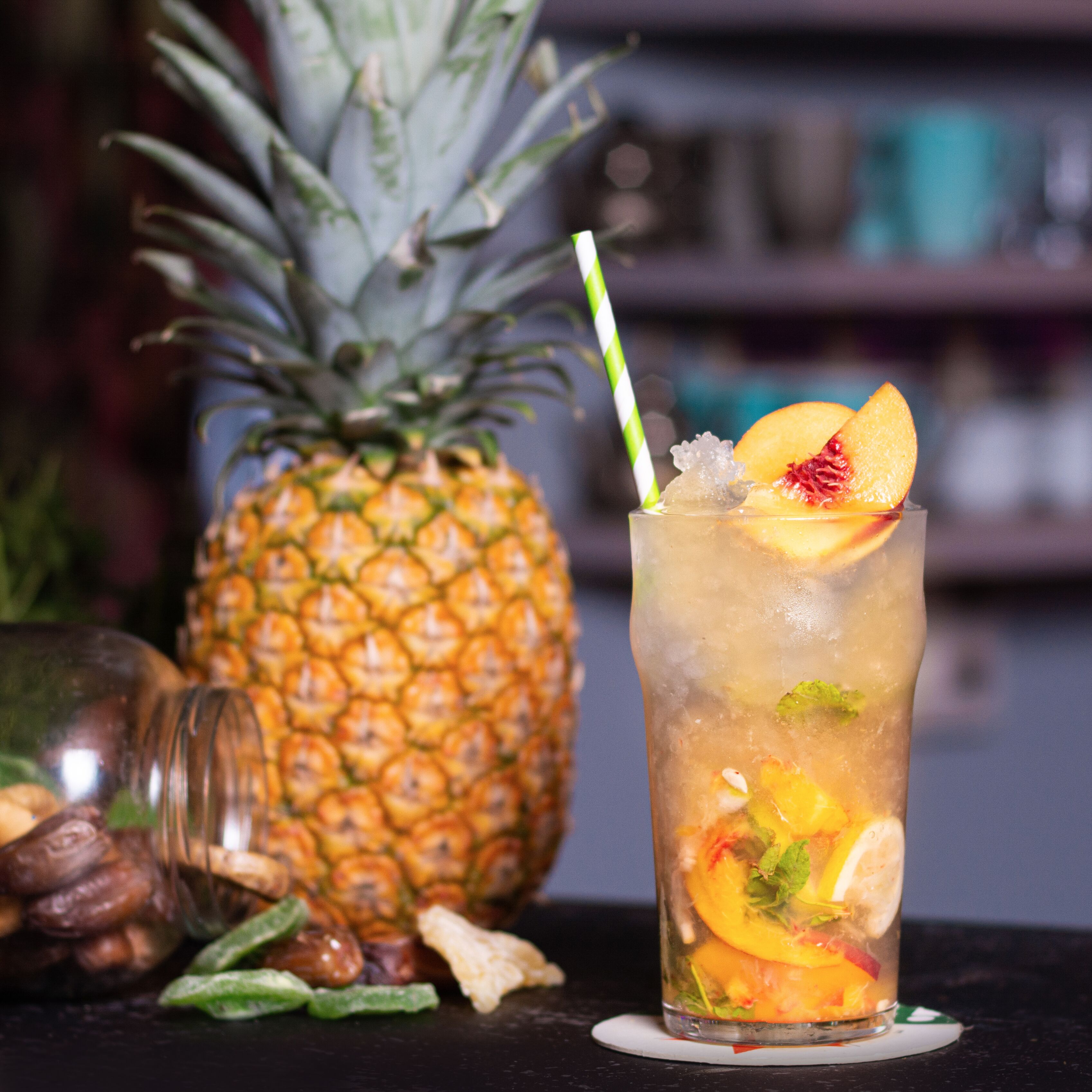 A glass of iced drink with fruit and mint, a pineapple, and a bottle of liquor on a dark surface.