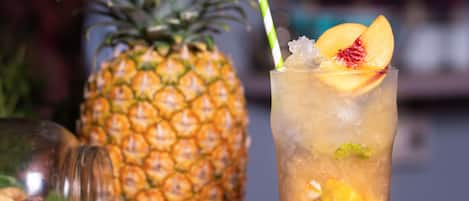 Um copo de bebida gelada com frutas e hortelã, um abacaxi e uma garrafa de licor sobre uma superfície escura.