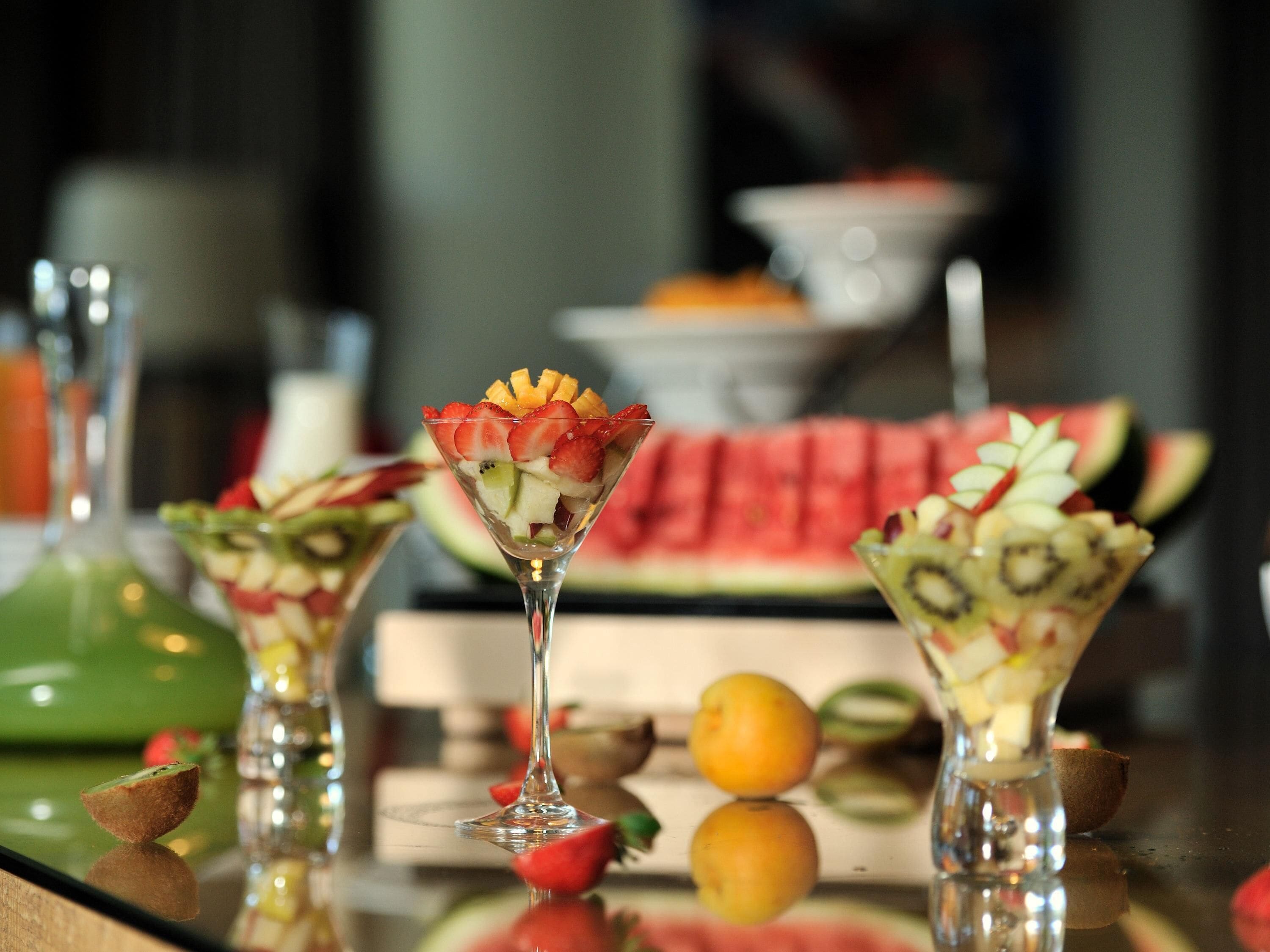 Uma mesa com três bebidas à base de fruta em copos de martini, uma melancia e uma garrafa verde.