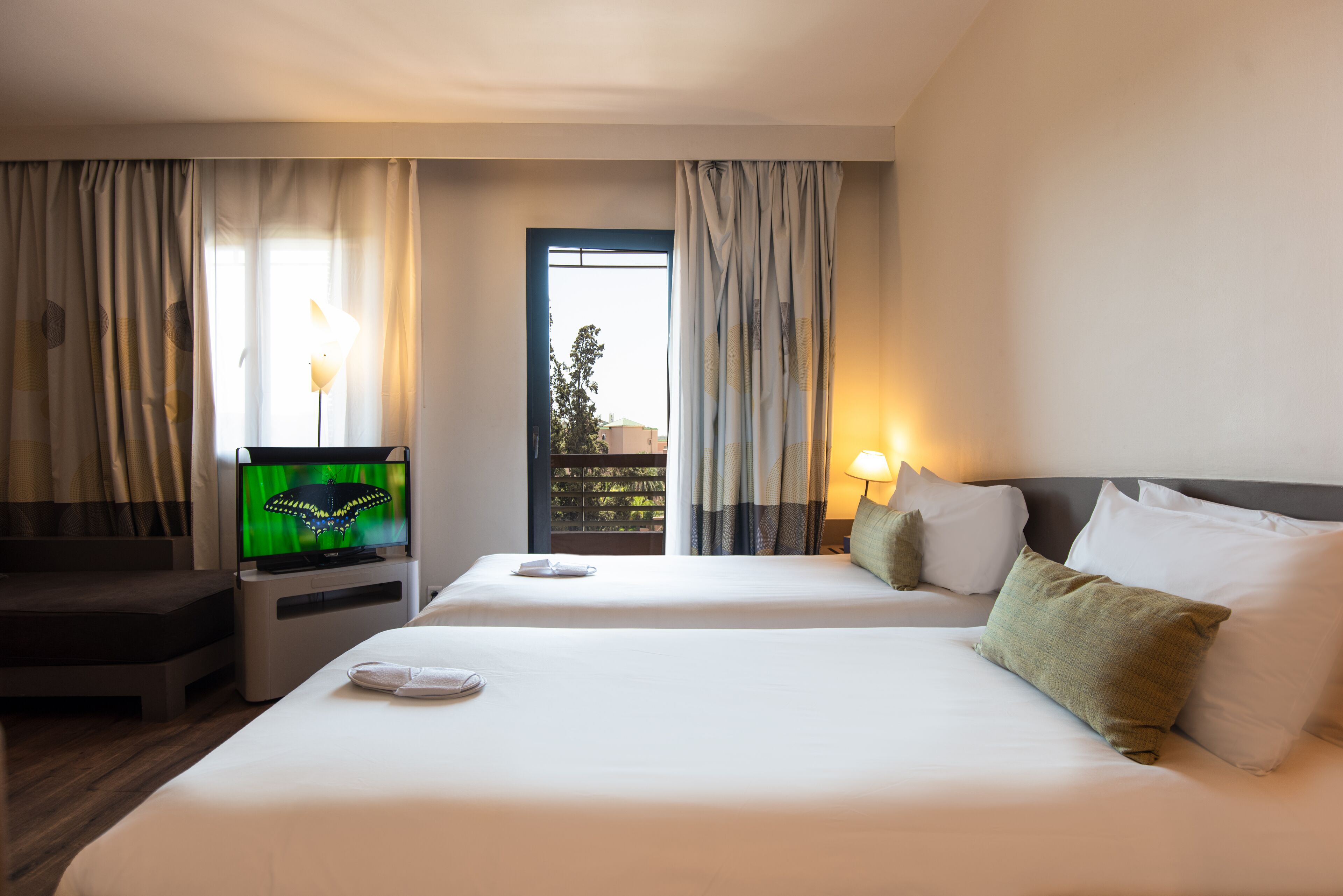 Quarto de hotel com duas camas, televisão, janela com cortinas e vista para o verde lá fora.